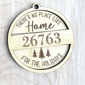 Puede incluir: Un adorno redondo de madera con el texto "There's no place like home 26763 for the holidays" y tres pinos estilizados.