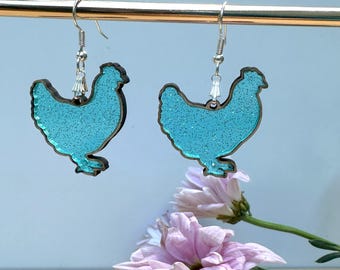 May include: A pair of dangle earrings featuring a turquoise glitter chicken design with a dark brown outline. The earrings hang from silver-colored hooks and are displayed against a light blue background with pink flowers.