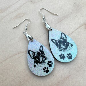 May include: A pair of iridescent blue teardrop-shaped earrings with a black and white image of a French bulldog and paw prints.