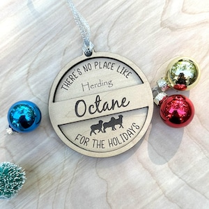 May include: A round wooden bauble with the text "There's no place like herding Octane for the holidays". The bauble is decorated with three sheep silhouettes. The bauble is hanging from a white ribbon and is surrounded by three colourful baubles.