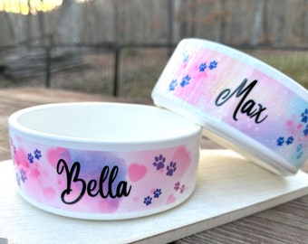 May include: Two white ceramic pet bowls with watercolor designs. One bowl is labeled "Bella" and the other "Max" in black script. Both bowls feature pink and purple hearts, paw prints, and a pastel background.