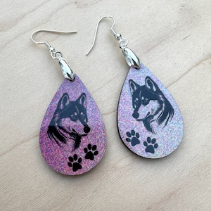 May include: A pair of teardrop-shaped earrings with a black and white husky silhouette and paw prints on a pink iridescent glitter background.