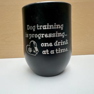 May include: Black stainless steel tumbler with the phrase "Dog training is progressing... one drink at a time." A heart-shaped graphic includes a paw print and a bottle. The tumbler is designed for beverages.
