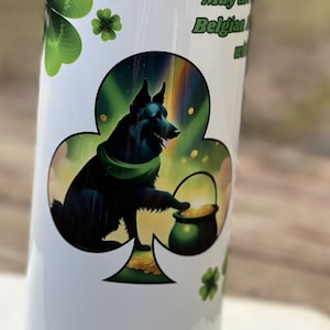 May include: A white tumbler with a green shamrock design. A black dog with a green scarf is holding a pot of gold. The text "May the Belgian Malinois" is visible on the tumbler.