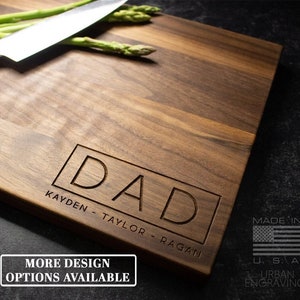 May include: A personalized walnut wood cutting board with the word "DAD" engraved in a rectangle. The board also has the names "KAYDEN - TAYLOR - RAGAN" engraved below the word "DAD". The board is made in the USA and has a small American flag graphic.