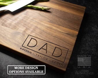May include: A personalized walnut wood cutting board with the word "DAD" engraved in a rectangle. The board also has the names "KAYDEN - TAYLOR - RAGAN" engraved below the word "DAD". The board is made in the USA and has a small American flag graphic.