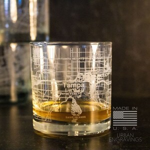 May include: A clear glass with a detailed etched map of Tampa, Florida. The glass is filled with a golden liquid and has the words "Tampa" and "Davis Islands" etched on the bottom. The glass is made in the USA by Urban Engravings.