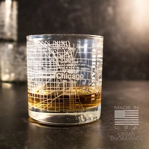 May include: A clear glass tumbler with an etched map of Chicago. The glass is filled with a brown liquid and has the words "Chicago" and "Made in U.S.A." on it. The glass is sitting on a black surface.
