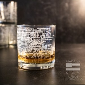 May include: A clear glass with a detailed etched map of Detroit, Michigan. The glass is filled with a brown liquid and has the text "MADE IN U.S.A. URBAN ENGRAVINGS" on the bottom.
