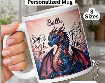 May include: A white ceramic mug featuring a blue and red dragon illustration with floral accents. The text reads Bella, Stay Soft at Heart, and Fierce When it Matters. The mug includes labels for Red & Blue Dragon Personalized Mug, 3 Sizes, and Add Name.