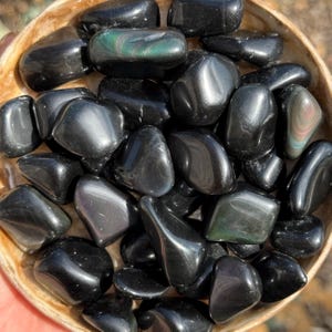 May include: A collection of polished black obsidian stones with iridescent patches, displayed in a light brown wooden bowl. The stones vary in size and shape, reflecting light and showcasing natural patterns. The overall aesthetic is smooth and organic.