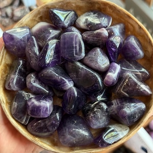 May include: A wooden bowl filled with polished amethyst stones. The stones vary in size and shape, displaying shades of purple and white. The bowl has a natural, light brown color.