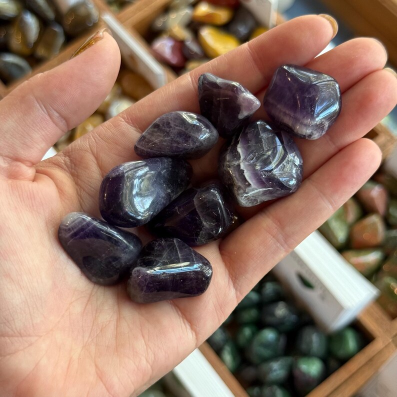 May include: A handful of polished amethyst stones, showcasing a deep purple hue with lighter, translucent streaks. The stones are smooth and rounded, reflecting light. The background includes other stones in wooden trays.