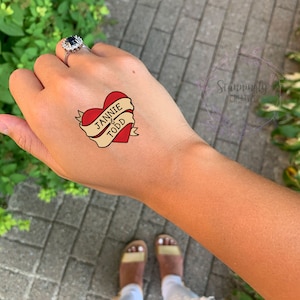 Puede incluir: Un tatuaje temporal en forma de coraz&oacute;n rojo con una pancarta que dice "Jannie & Todd".