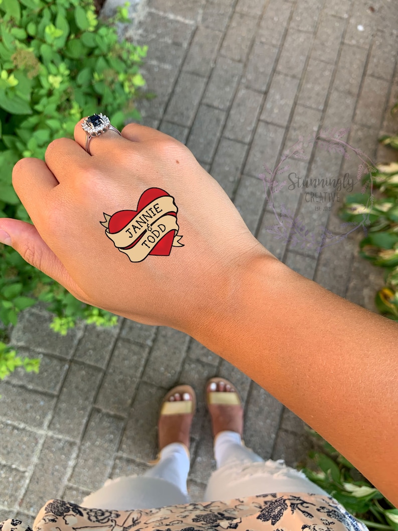 Puede incluir: Un tatuaje temporal en forma de coraz&oacute;n rojo con una pancarta que dice "Jannie & Todd".