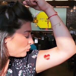 May include: A woman with a red string bracelet on her wrist has a small red heart tattoo on her upper arm with the word "Nicky" written inside the heart.