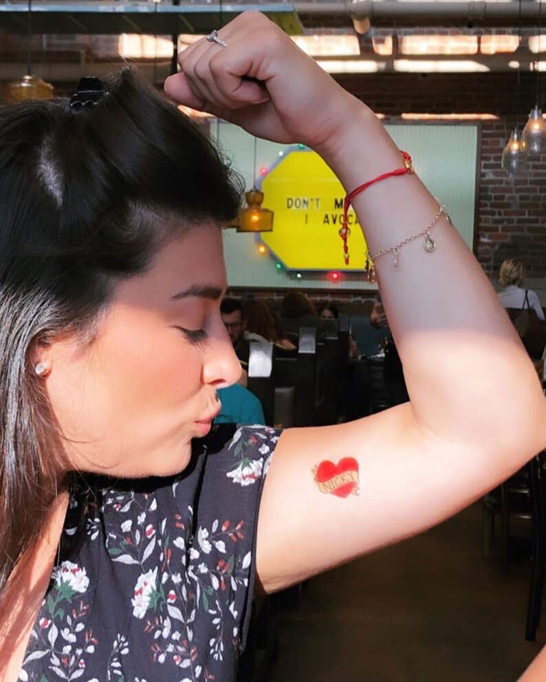 May include: A woman with a red string bracelet on her wrist has a small red heart tattoo on her upper arm with the word "Nicky" written inside the heart.