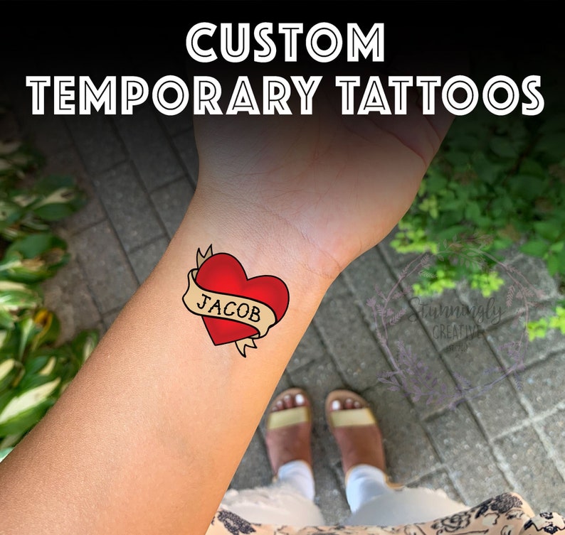 May include: A red heart with a banner that says "JACOB" on a temporary tattoo. The tattoo is on a person's wrist.