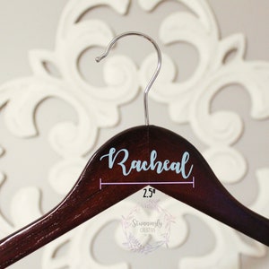 May include: A dark brown wooden hanger with a light blue vinyl name "Rachel" printed on the top. The hanger is 2.5 inches wide.