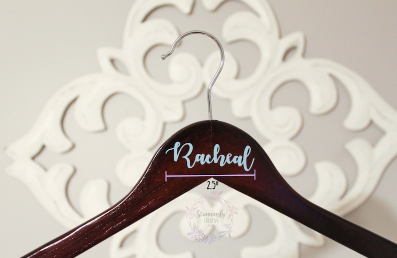 May include: A dark brown wooden hanger with a light blue vinyl name "Rachel" printed on the top. The hanger is 2.5 inches wide.