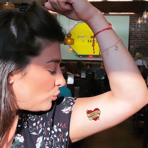 Puede incluir: Un tatuaje de coraz&oacute;n rojo con una pancarta que dice "Danielle Nicky" en el brazo de una persona.