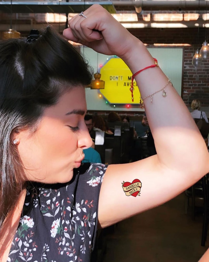 Puede incluir: Un tatuaje de coraz&oacute;n rojo con una pancarta que dice "Danielle Nicky" en el brazo de una persona.