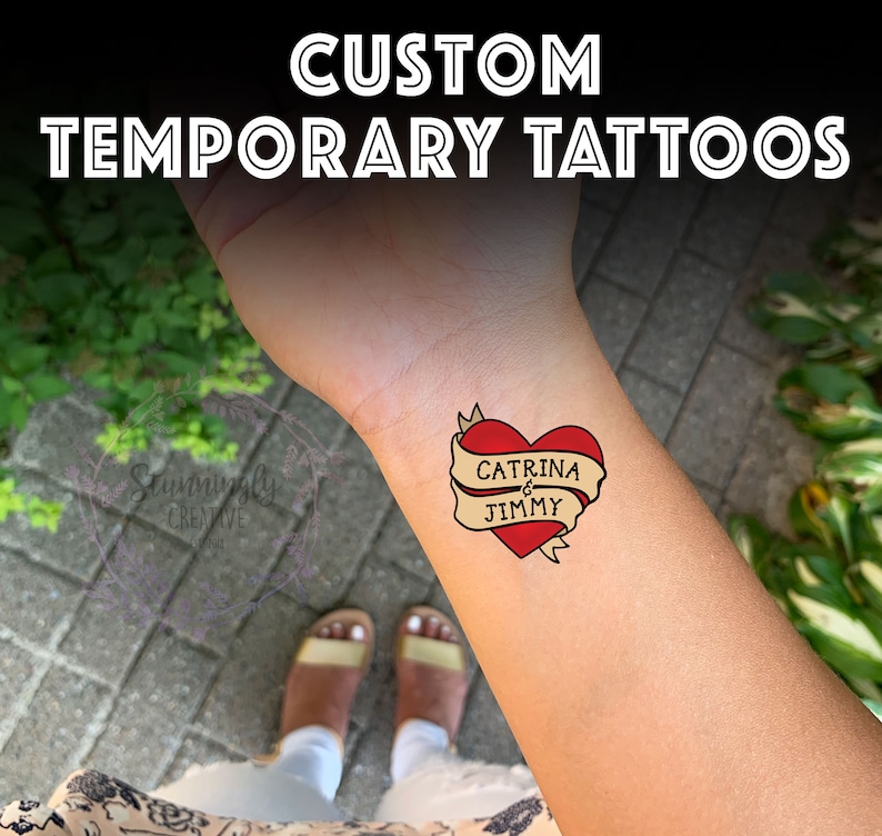 Puede incluir: Un tatuaje temporal con un coraz&oacute;n rojo y una pancarta amarilla que dice "CATRINA & JIMMY".