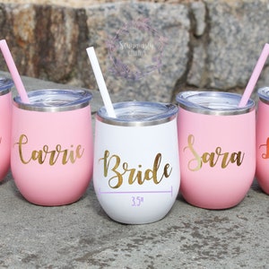 May include: Five pink and white stainless steel wine tumblers with gold lettering. The tumblers are labeled "Stormi", "Carrie", "Bride", "Sara", and "Le Land". Each tumbler has a straw.