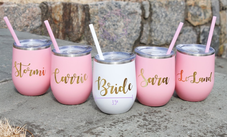 May include: Five pink and white stainless steel wine tumblers with gold lettering. The tumblers are labeled "Stormi", "Carrie", "Bride", "Sara", and "Le Land". Each tumbler has a straw.