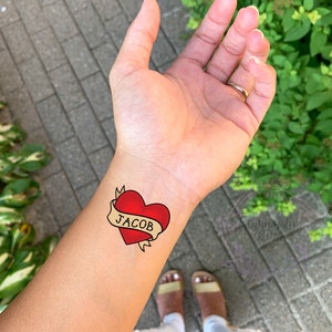 May include: A red heart with a yellow banner that says "JACOB" on it. The heart is a temporary tattoo.