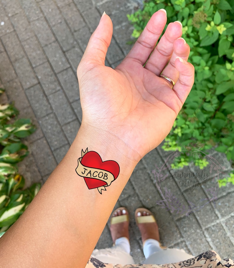 May include: A red heart with a yellow banner that says "JACOB" on it. The heart is a temporary tattoo.