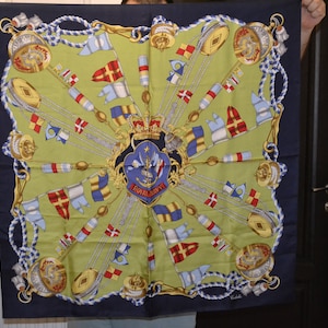 May include: A green and blue silk scarf with a nautical theme. The centre of the scarf features a crest with a crown, an anchor, and the words "Royal Navy". The scarf is surrounded by a border of ropes and nautical flags.