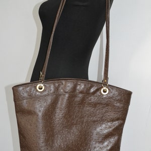 Può includere: Una borsa tote in pelle marrone con un motivo testurizzato simile a quello di struzzo. La borsa ha due manici superiori e una lunga tracolla.