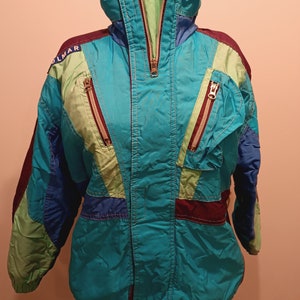 Peut inclure: Une veste de ski vintage dans un schéma de couleurs bleu vif, vert et violet avec une fermeture à glissière et plusieurs poches. La veste porte le mot "OLMAR" sur le côté gauche de la poitrine.