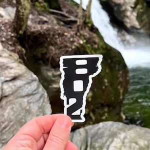 May include: A black and white sticker of the state of Vermont with the number 802 inside. The sticker is being held in front of a waterfall.
