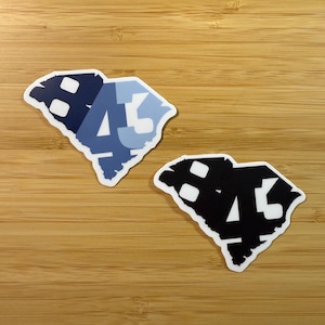 May include: Two stickers featuring the outline of the state of South Carolina. One sticker is in shades of blue with the numbers "45" overlaid. The other sticker is black with white accents and the same "45" design.