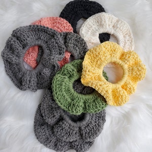 May include: A set of nine crocheted hair scrunchies in various colors, including gray, black, white, yellow, green, and pink. The scrunchies are arranged in a pile on a white fluffy surface.