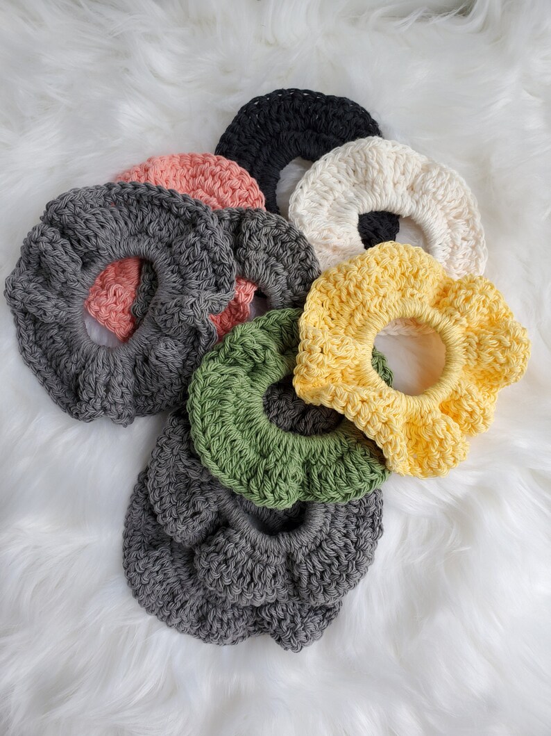 May include: A set of nine crocheted hair scrunchies in various colors, including gray, black, white, yellow, green, and pink. The scrunchies are arranged in a pile on a white fluffy surface.