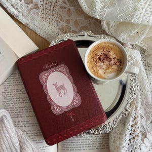 May include: A dark red book with the word Bookish and a deer silhouette on the cover, placed next to a white ceramic cup filled with a foamy coffee drink on a silver tray, all resting on an open book with visible text and white lace fabric.