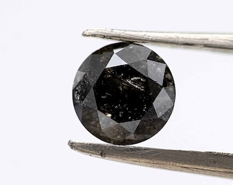 May include: A black round diamond with a faceted cut, held by tweezers. The diamond has a dark gray color and a rough, textured surface.