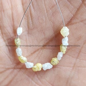 May include: A strand of raw, uncut diamonds in shades of white and yellow. The rough gemstones are strung together on a thin wire, showcasing their natural, irregular shapes. The image is taken close-up, highlighting the texture and color variations of the diamonds.