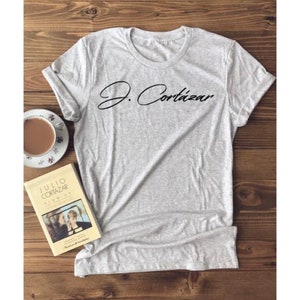 Könnte beinhalten: Ein graues T-Shirt mit dem Text "J. Cortázar" in schwarzer Schrift auf der Vorderseite. Im Hintergrund ist ein Buch mit dem Titel "Julio Cortázar Blow Up" zu sehen.