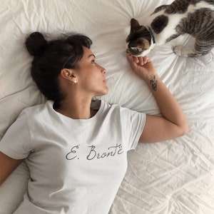 Puede incluir: Una mujer lleva una camiseta blanca con el texto "E. Bront&euml;" en escritura negra. Est&aacute; tumbada en una cama blanca con un gato a su lado.