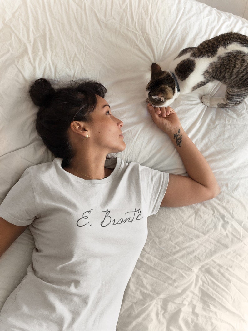 Puede incluir: Una mujer lleva una camiseta blanca con el texto "E. Bront&euml;" en escritura negra. Est&aacute; tumbada en una cama blanca con un gato a su lado.