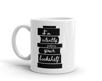 Puede incluir: Taza de café de cerámica blanca con un gráfico negro de una pila de libros que dice "Estoy juzgando silenciosamente tu estantería".