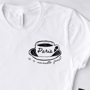 May include: White t-shirt with a black graphic of a coffee cup and saucer. The word "Paris" is written inside the cup, and the phrase "is a moveable feast" is below the saucer. The shirt is made of a soft material.