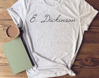 Può includere: Una maglietta grigia con il testo "E. Dickinson" stampato in lettere corsive nere.