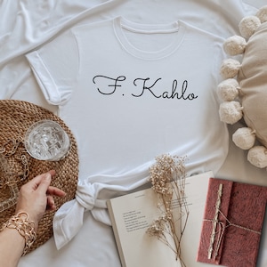 Könnte beinhalten: Weißes T-Shirt mit schwarzem Text, der "F. Kahlo" auf weißem Hintergrund lautet. Das Shirt liegt auf einer weißen Oberfläche mit einem Buch, einem Glas Eis und einem Dekokissen.