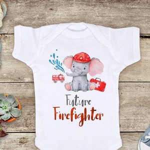 May include: White baby onesie with a watercolor illustration of an elephant wearing a firefighter hat and holding a fire hose. The text "Future Firefighter" is printed below the elephant.