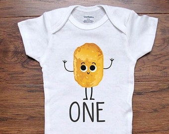 May include: White baby onesie with a cartoon illustration of a golden brown nugget with a smiling face and arms and legs. The text "ONE" is printed below the nugget.